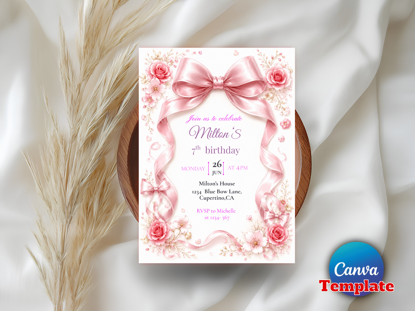 A pink floral birthday invitation card with a large bow illustration, styled on a white satin fabric with dried pampas grass and a wooden tray. A blue circular 'Canva Template' badge appears in the bottom right corner.